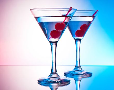 Two glasses of martini with red cherries on a mixed color background