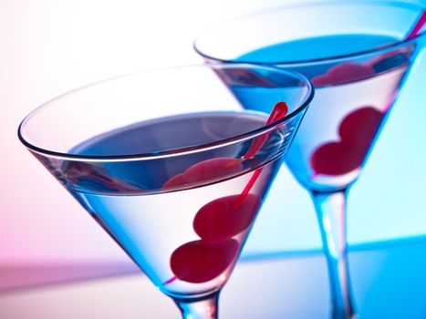 Two glasses of martini with red cherries on a mixed color background