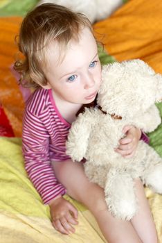 Sad little girl with blue eyes sitting on a bed with a teddy bear.