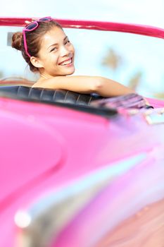 Retro woman smiling happy in old pink vintage car driving on road trip on beautiful summer day. Pretty mulitracial Asian / Caucasian female model in Havana, Cuba