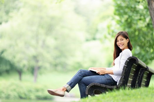Park woman reading book on bench smiling happy at camera. Pretty young multicultural woman enjoying spring in park.