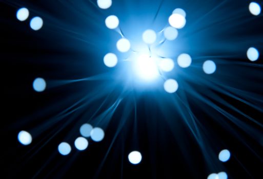 Photograph capturing the blurred ends of many illuminated blue fiber optic strands emerging from a central ilght source with black background.