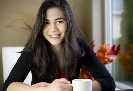 Beautiful biracial teenage girl at table with coffee mug in hand, smiling