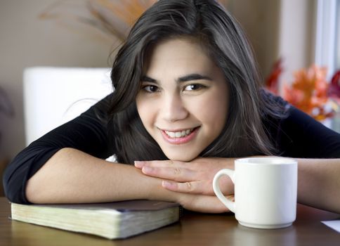 Beautiful biracial teenage girl at table with chin on arm, relaxing with a cup of coffee and her Bible or book 