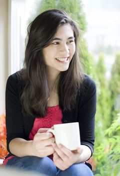 Beautiful biracial teenager or young woman drinking coffee next to window