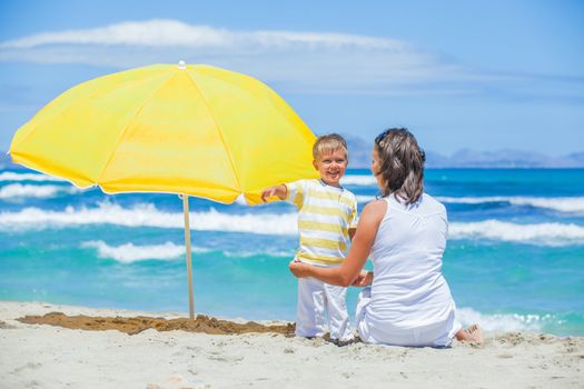 Back view of cute 2 years old boy and his mother with big umbrella on tropical beach