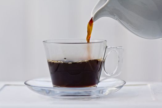 Pouring coffee into a half full clear cup with a saucer.