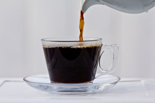 Pouring coffee into an almost full clear cup with a saucer.