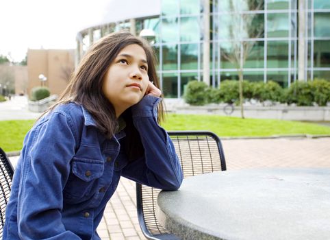 Twelve year old girl sitting at outdoors table looking up into sky with thoughtful expression, head on hand