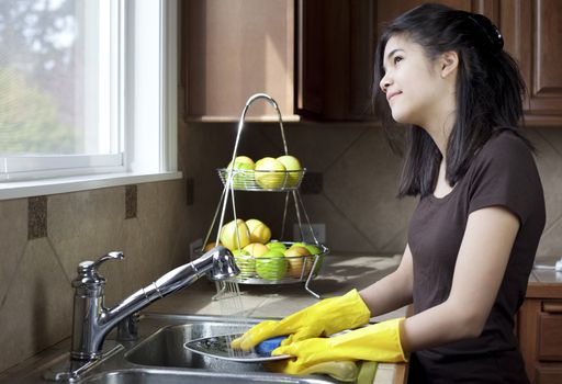 Teen girl washing dishes at kitchen sink, daydreaming or looking out the window with thoughtful expression.