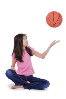 Ten year old Asian girl sitting on floor throwing basketball up in the air, isolated on white