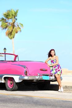 Vintage car and woman standing happy and smiling leaning on pink retro car on the side of the road. Beautiful young multicultural young woman on spring or summer road trip. Havana, Cuba.