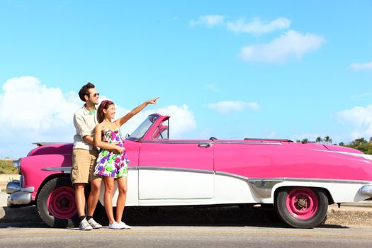Vintage car. Couple pointing looking standing by pink retro vintage car smiling happy. Young couple on summer road trip car holiday in Havana, Cuba. Asian woman, Caucasian man.