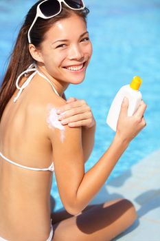 Sunscreen woman putting solar cream or sunblock smiling happy in bikini by pool. Beautiful young woman under the sun on sunny summer day, Mixed race Asian / Caucasian girl.