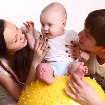 Portrait of a young family on a white background. The father, mum and the son. A square format. The kid sits on a yellow ball, parents hold the kid