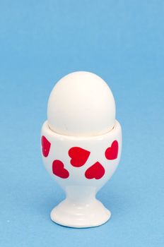 white egg in a special cup for eating covered with red hearts on blue background isolated.