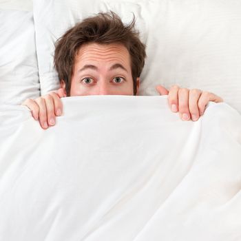 Scared man hiding in bed under the sheets. Funny concept image with young caucasian male model home in bed.