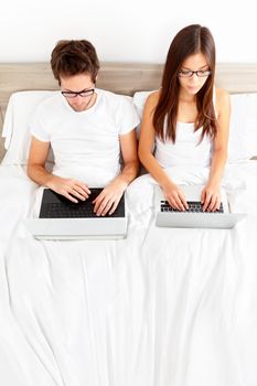 Couple on laptop in bed working on separate computers. Young modern interracial couple, Asian woman, Caucasian man in high angle view with copy space.