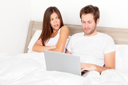 Couple with laptop in bed. Man at computer, woman upset and angry looking at man. Young modern interracial couple in bed. Asian woman, Caucasian man. Funny image.