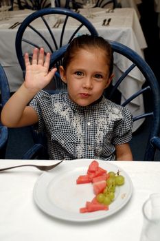 A sweet child sitting at table in a restaurant waving with her hand