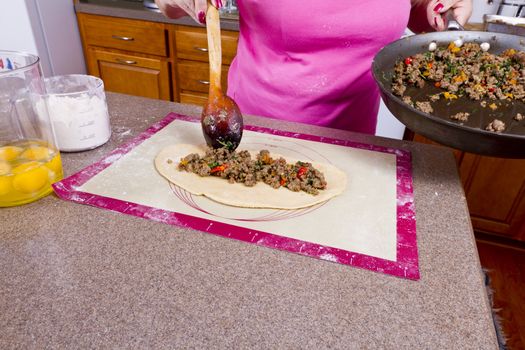 Well used wooden utensil putting, cooked seasoned ground beef on to dough to make Turkish Pide pocket.