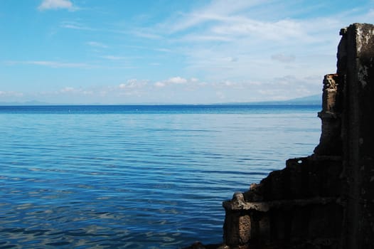 blue water bay and blue sky seen beyond ruins of concrete wall