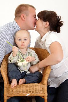 Mother and father are kissing while their baby is playing with a flower .