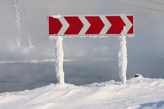 Frozen and snowed guide road sign of turn on a background of a fog above water