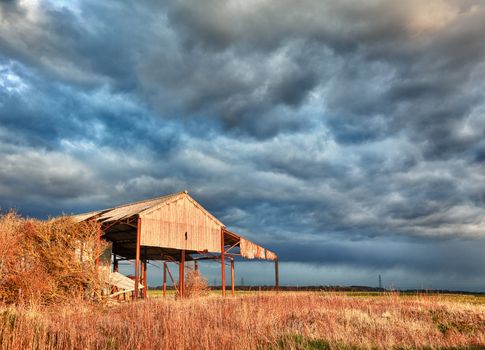 Storm clouds over sunlit deserted and ruined barn on farmland