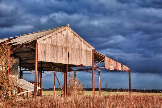 Storm clouds over sunlit deserted and ruined barn on farmland