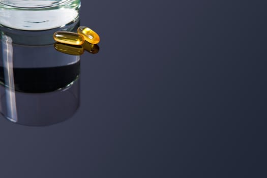 A couple of yellow vitamin pills on a black glass table with a glass of water at the side