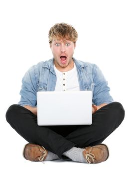 Laptop computer man shocked looking surprised at screen. Young modern male student model sitting cross legged on floor isolated on white background.