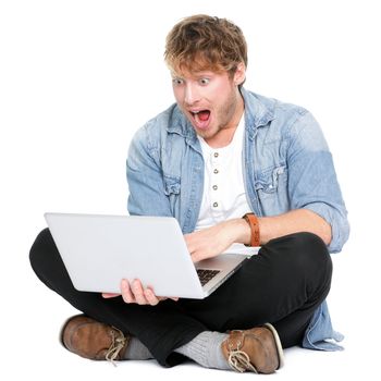 Man surprised with laptop computer looking at screen excited and happy in disbelief. Funny image of young Caucasian male student model sitting on floor isolated on white background.