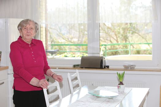 Elderly smiling lady standing next to a white table in front of large light windows