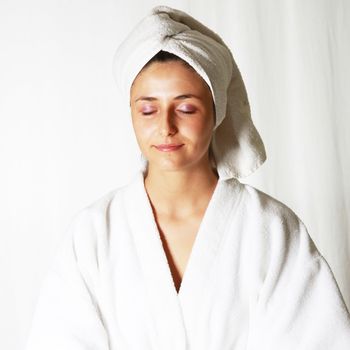 Headshot of a woman with her eyes closed and a serene look meditating in a bathrobe with a white towel wrapped around her head