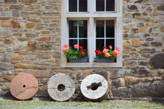 Rustic wooden sash window in an old stone walled building with colourful flower boxes of geraniums and round grindstones balanced on the grass below