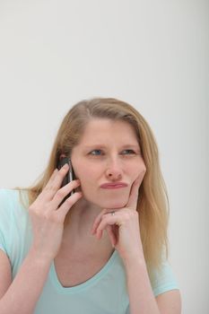 Sceptical young blonde woman sitting on her mobile phone with her chin on her finger and a look of disbelief and uncertainty