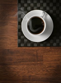 Steaming hot cup of black coffee on wooden table and dark tablemat