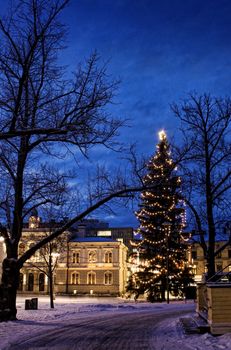 Illuminated tall Christmas tree in snowy old town centre evening