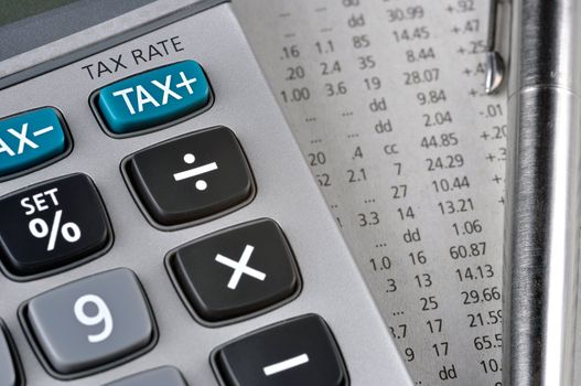 Detail of calculator, focusing the TAX key, next to a sheet of paper with numbers and a metal pen.