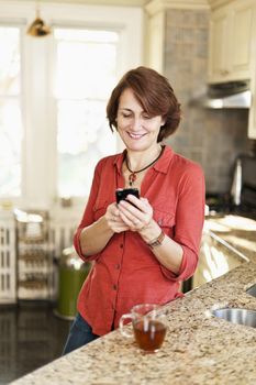 Smiling mature woman texting on phone in kitchen at home