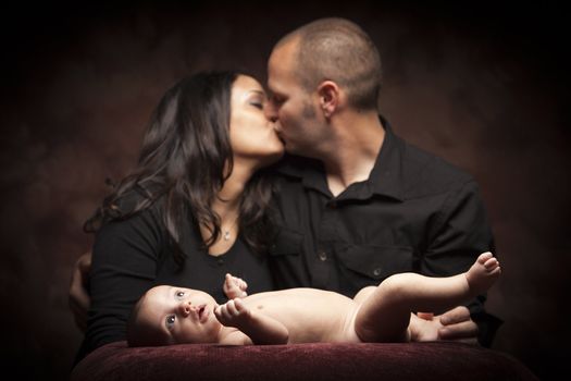 Mixed Race Couple Kiss While Baby Lays on Pillow on a Dark Background.