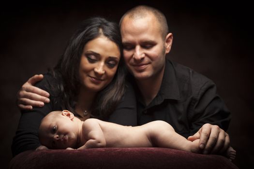 Mixed Race Couple Lovingly Look On While Baby Lays on Pillow on a Dark Background.