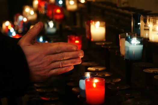 Religion concept: Praying hands in front of a series of colorful glass church candle at a place of worship; soft candlelight lighting.
