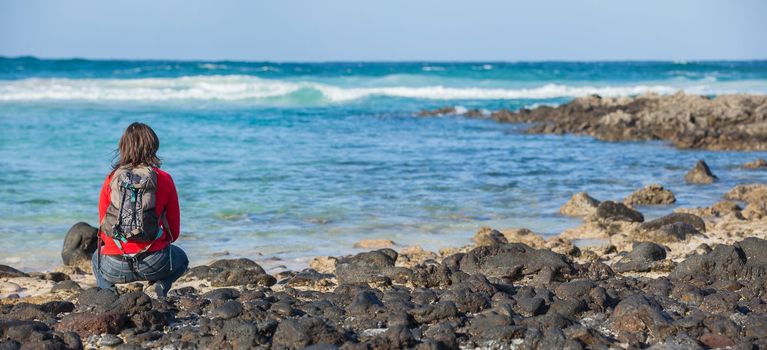 Back view of sports young woman with backpack sits on the beach and looks at the sea