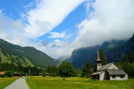 Small Church near Path in Meadow with Mountains in Background and Cloudy Sky taken in Kandersteg, Switzerland. Landscape Orientation.