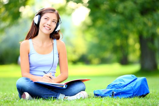 Female student girl outside in park listening to music on headphones while studying. Happy young university student of mixed Asian and Caucasian ethnicity.