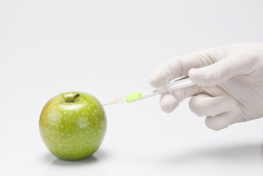 A gloved hand injecting an apple with a syringe on a white background.