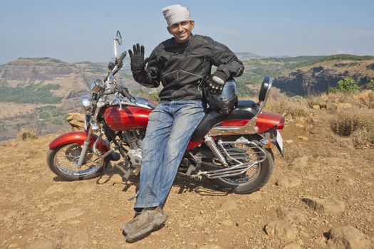 Motorbiker with a red cruiser relaxing on reaching the top of a hill at Tiger Point, Maharastra, India, waving to the camera