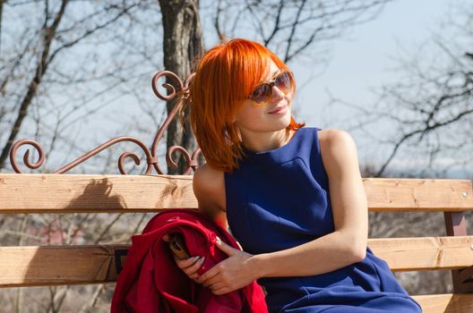 Beautiful red woman wearing sunglasses sitting on the bench on a sunny day